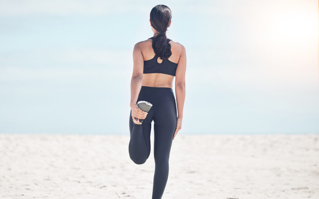 Fitness, woman back and stretching leg at beach for running, workout and body, exercise or wellness routine. Foot, stretch and behind female runner at sea for training, sports or physical performance